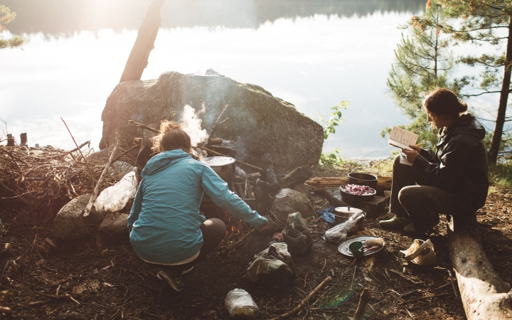 On the shore of a lake, one person tends to a campfire, while another sits nearby and reads. 